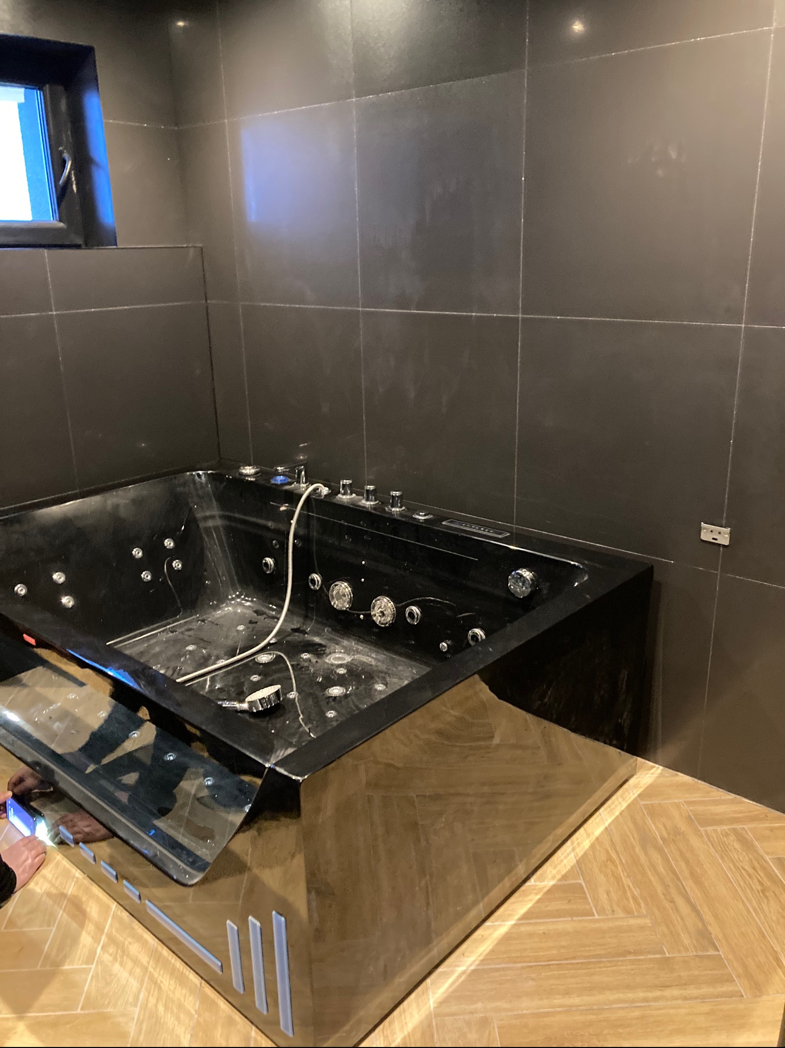 Czarna wanna z hydromasażem, z chromowanymi elementami sterującymi i dyszami, zamontowana w łazience z ciemnymi, kwadratowymi płytkami, częściowo widoczna podłoga z jasnego drewna ułożonego w jodełkę.