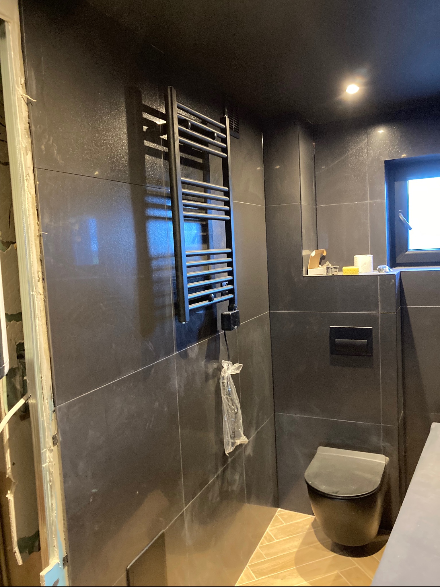 Ciemna łazienka w trakcie urządzania: czarne płytki, grzejnik drabinkowy, wisząca toaleta i fragment blatu umywalki. Widoczne niedokończone obramowanie drzwi i akcesoria na półce.