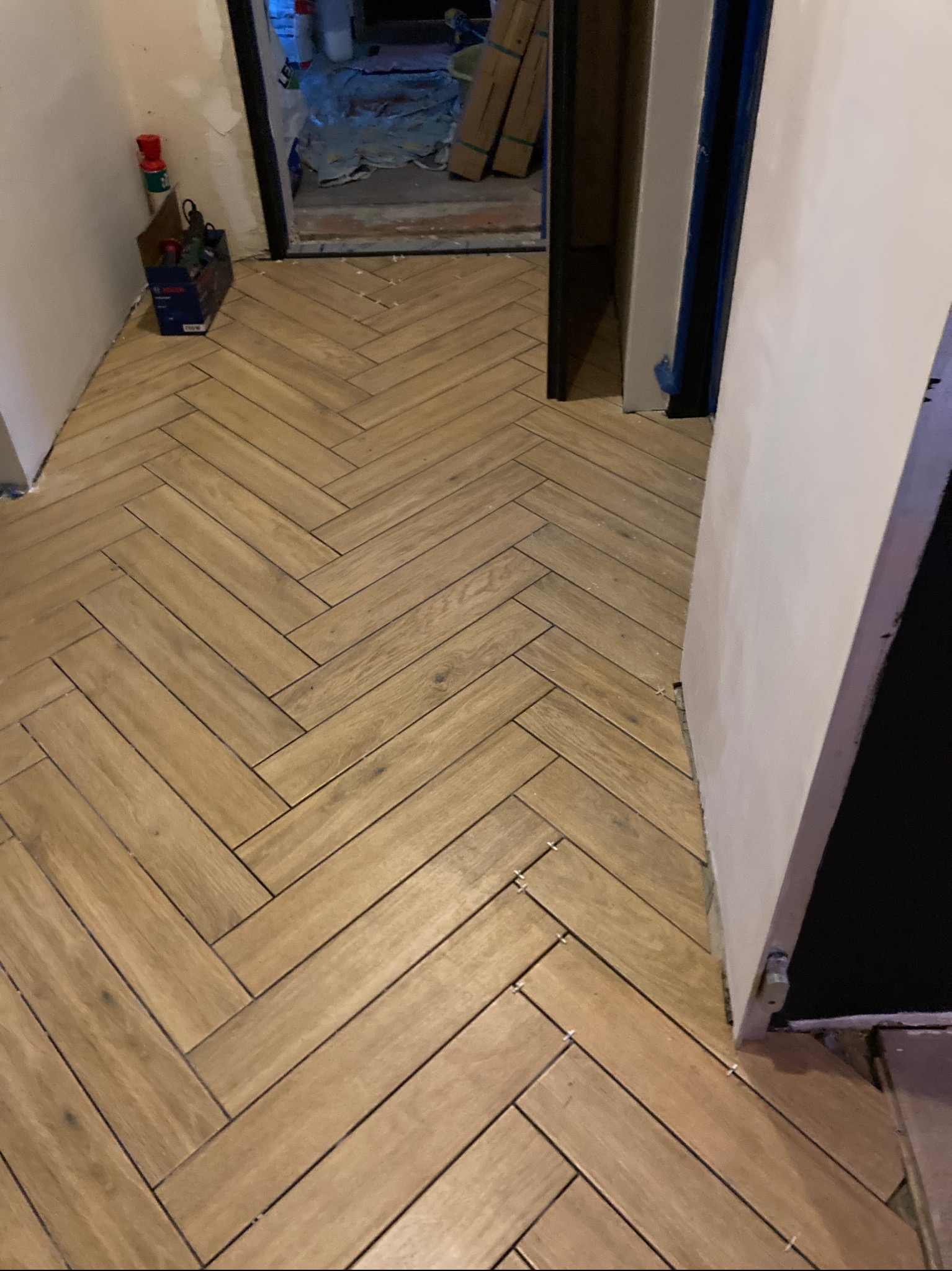 Ułożona podłoga z płytek imitujących drewno w jodełkę francuską, widoczne krzyżyki dystansowe, w tle narzędzia i materiały budowlane.