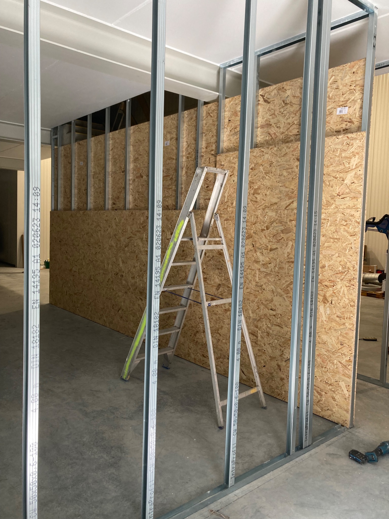 Konstrukcja ścian działowych z profili stalowych i płyt OSB, widoczna drabina aluminiowa i wkrętarka akumulatorowa na posadzce.