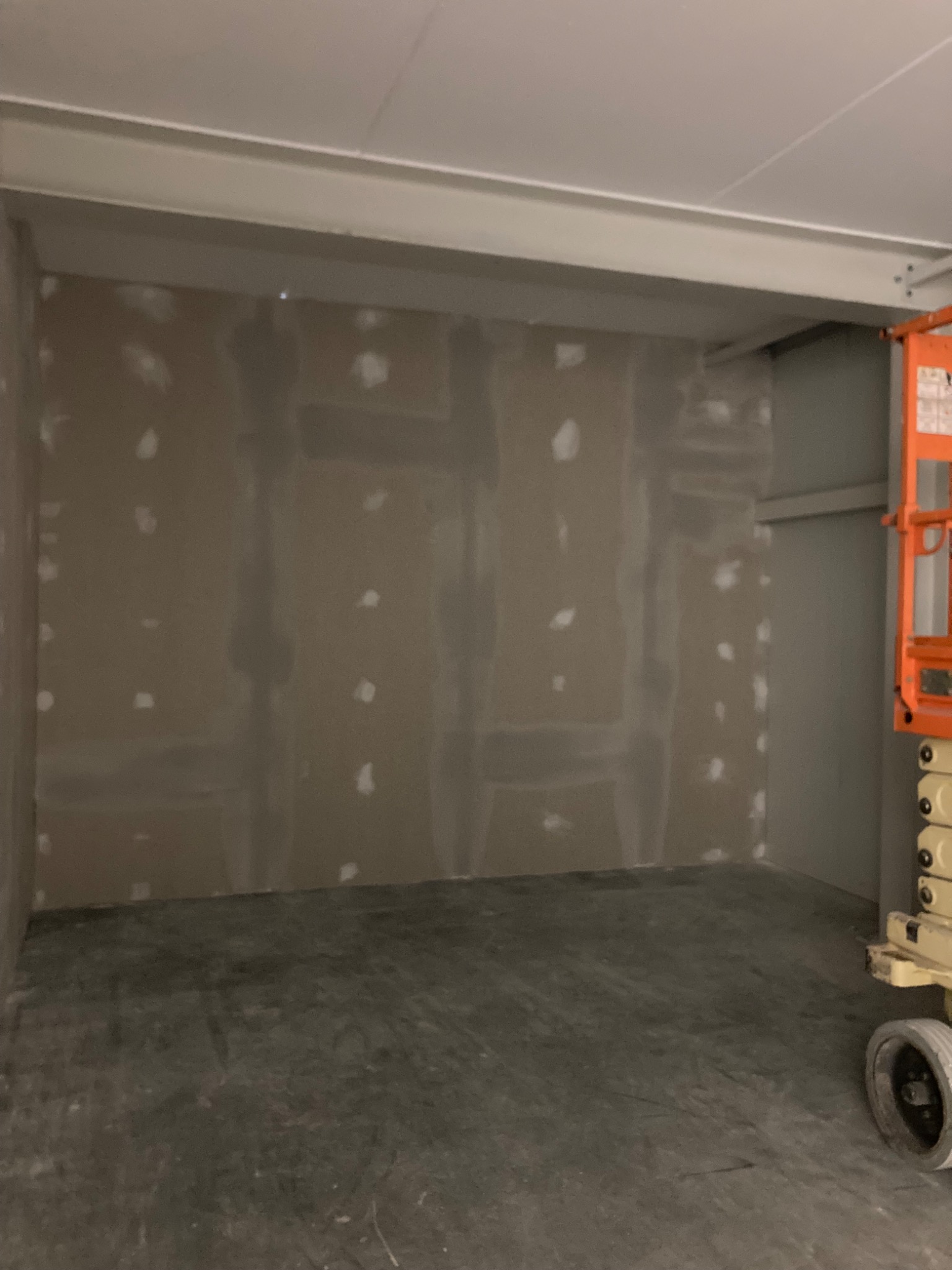 Przygotowana ściana z płyt gipsowo-kartonowych przed nałożeniem gładzi, widoczne miejsca po wkrętach pokryte masą szpachlową, na podłodze ślady po pracach, w tle podnośnik nożycowy.