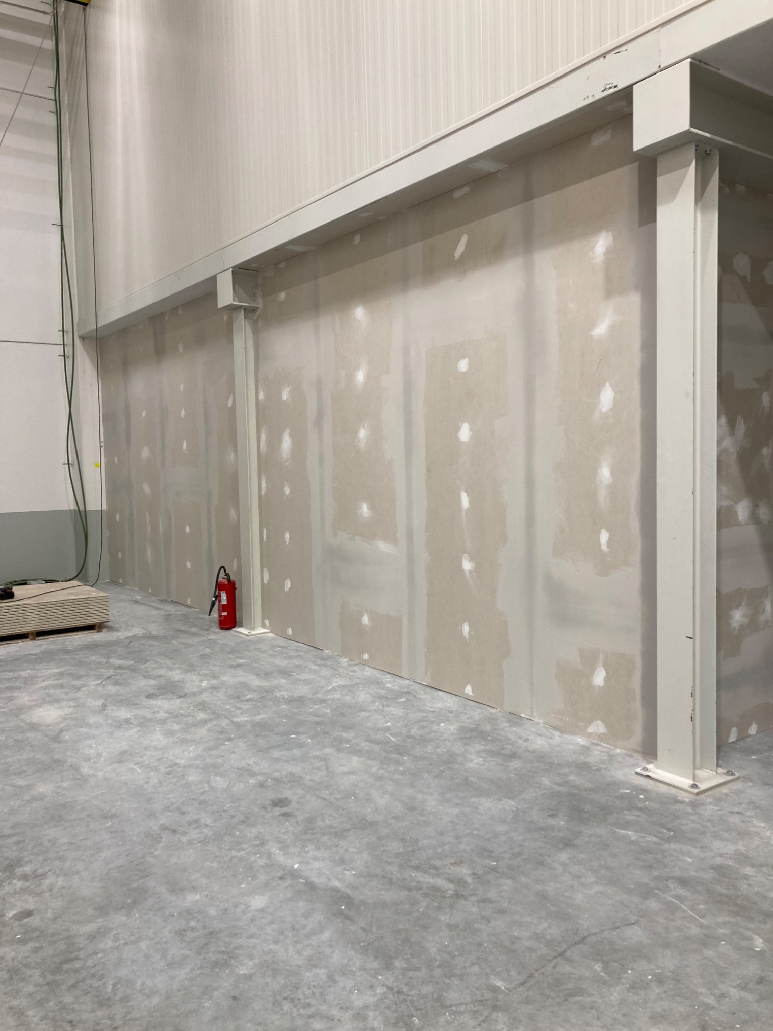 Ściana z płyt gipsowo-kartonowych w trakcie szpachlowania, widoczne białe punkty szpachli, czerwona gaśnica przy ścianie, przygotowane płyty gipsowe na podłodze w dużym, surowym pomieszczeniu.