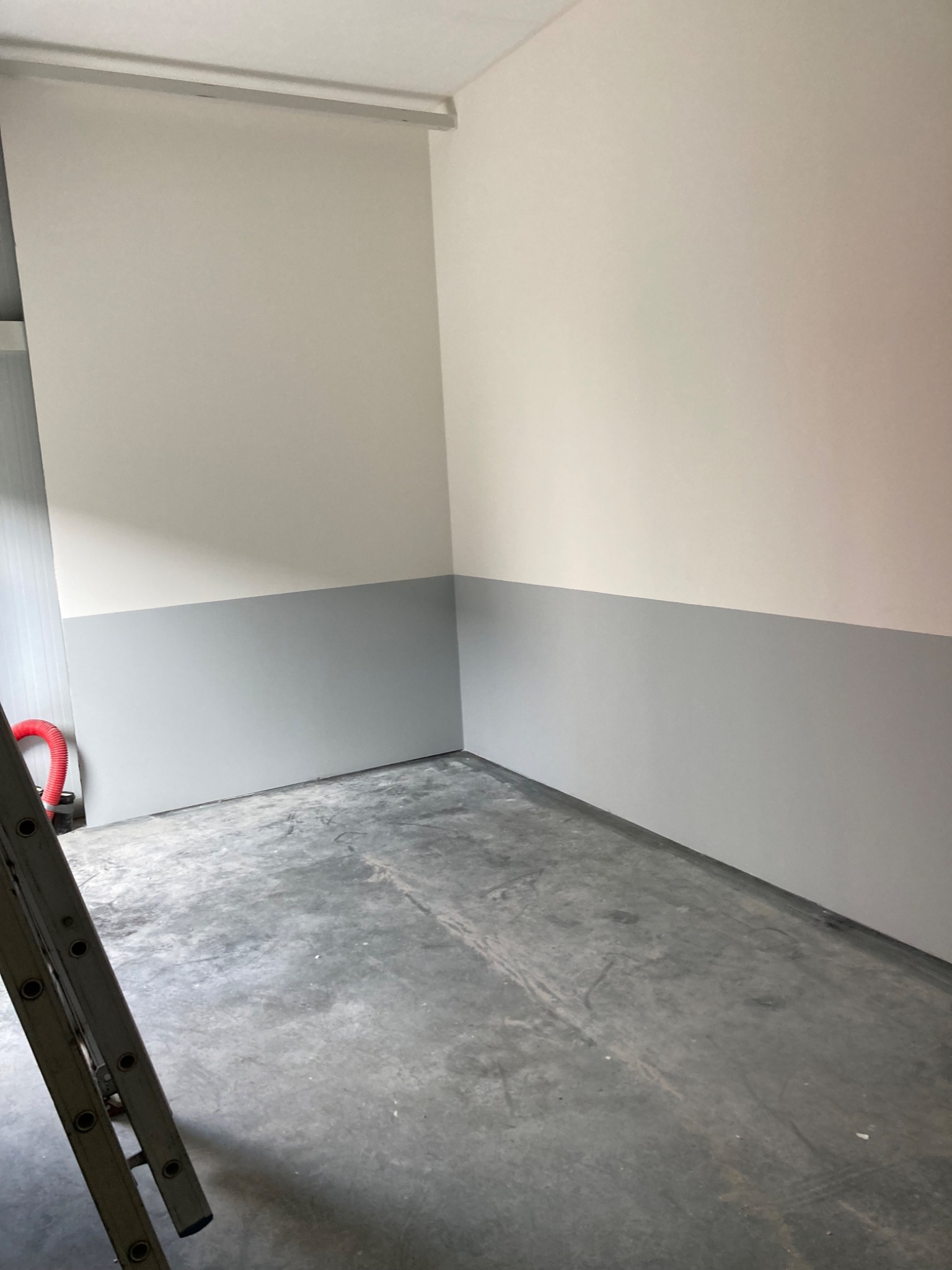 Pomieszczenie po malowaniu ścian na dwa kolory: górna część biała, dolna szara, widoczna drabina oparta o ścianę.