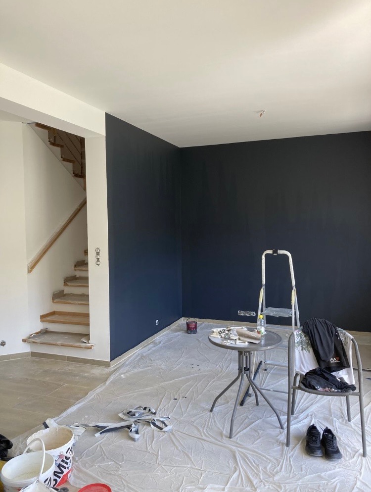 Pokój w trakcie malowania na ciemny granat, zabezpieczona podłoga folią, widoczna drabina, stolik z narzędziami i schody w tle.