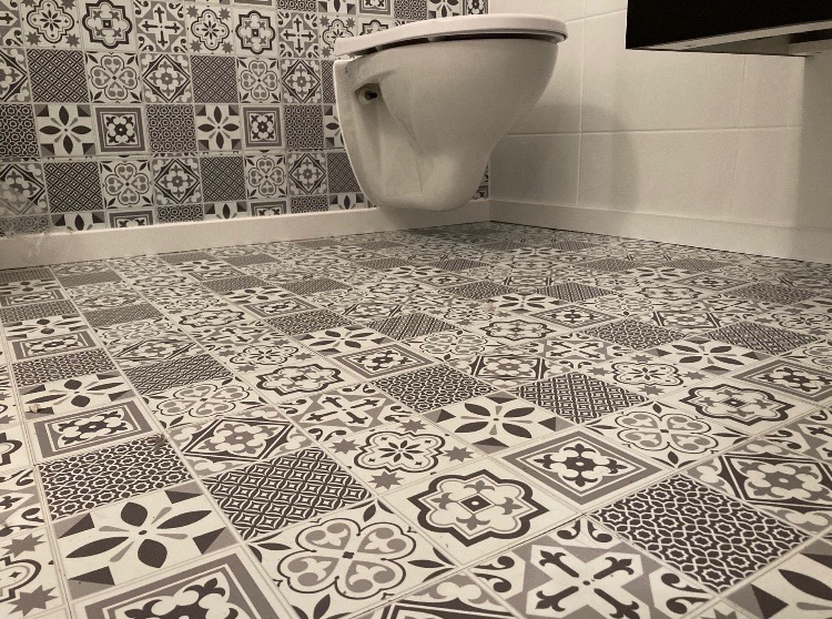 Ukończona łazienka z białą wiszącą toaletą i podłogą z wzorzystych, szaro-białych płytek, dopasowanych do płytek na ścianie za toaletą.