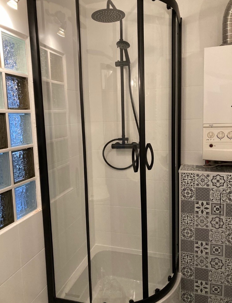 Kabina prysznicowa narożna z czarnymi profilami, białe płytki na ścianach, dekoracyjne luksfery i wzorzysta zabudowa obok kotła.