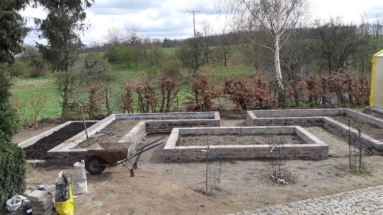 Budowa podwyższonych grządek warzywnych z cegły klinkierowej, częściowo wypełnionych ziemią, taczka i worki z ziemią w pobliżu, widok na ogród w tle.