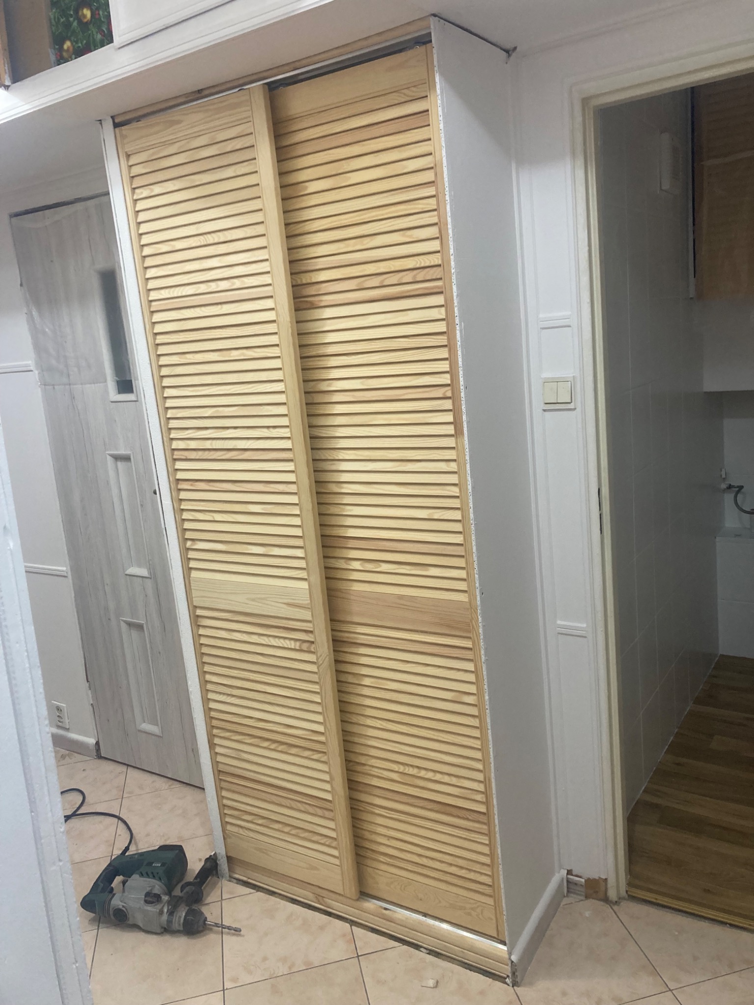 Montaż przesuwnych drzwi lamelowych w mieszkaniu, widoczny młot udarowy na podłodze, częściowo wykończona ściana i fragment łazienki w tle.