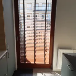 Brązowe poziome osłony okienne zamontowane w drewnianej ramie, widok na budynek z zewnątrz przez szpary, fragment kuchennej zabudowy po bokach.