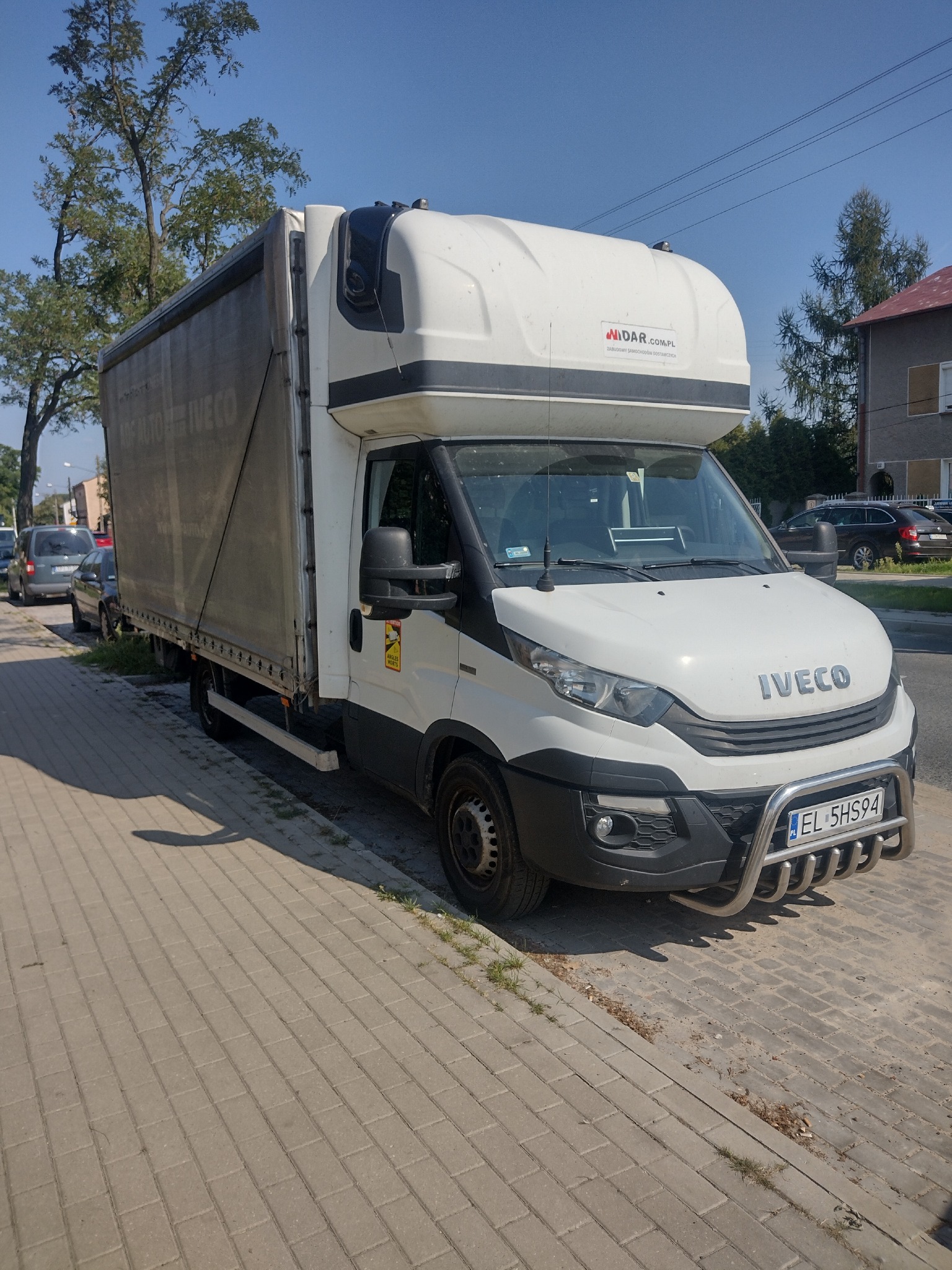 Biały samochód dostawczy Iveco z plandeką, zaparkowany na chodniku w słoneczny dzień, z widoczną tablicą rejestracyjną EL 5HS94 i logiem MIDAR.COM.PL na zabudowie.