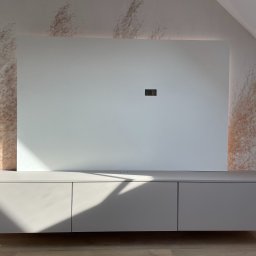 AVAR BESPOKE SPÓŁKA Z OGRANICZONĄ ODPOWIEDZIALNOŚCIĄ - Szara szafka RTV podwieszana pod białą ścianką z ukrytym oświetleniem LED, na tle ściany z wzorem trawy pampasowej, w pokoju na poddaszu.