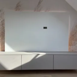 Szara szafka RTV podwieszana pod białą ścianką z ukrytym oświetleniem LED, na tle ściany z wzorem trawy pampasowej, w pokoju na poddaszu.