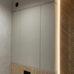 Beżowa szafka wisząca z trzema drzwiczkami, zamontowana w łazience z drewnianą boazerią i oświetleniem LED, widoczny przycisk spłuczki.