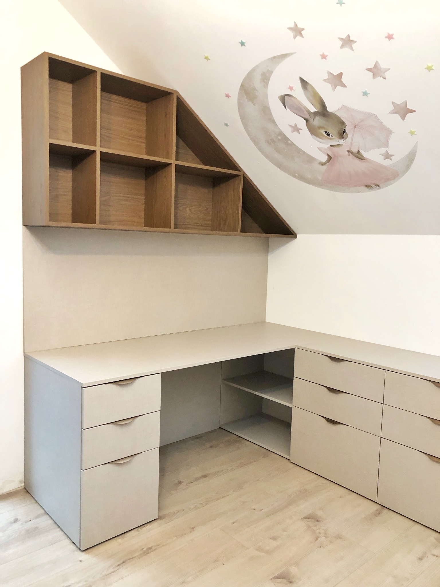 Biało-beżowe biurko narożne z szufladami i półkami, nad nim drewniany regał z przegródkami, na ścianie naklejka z księżycem i królikiem, poddasze.