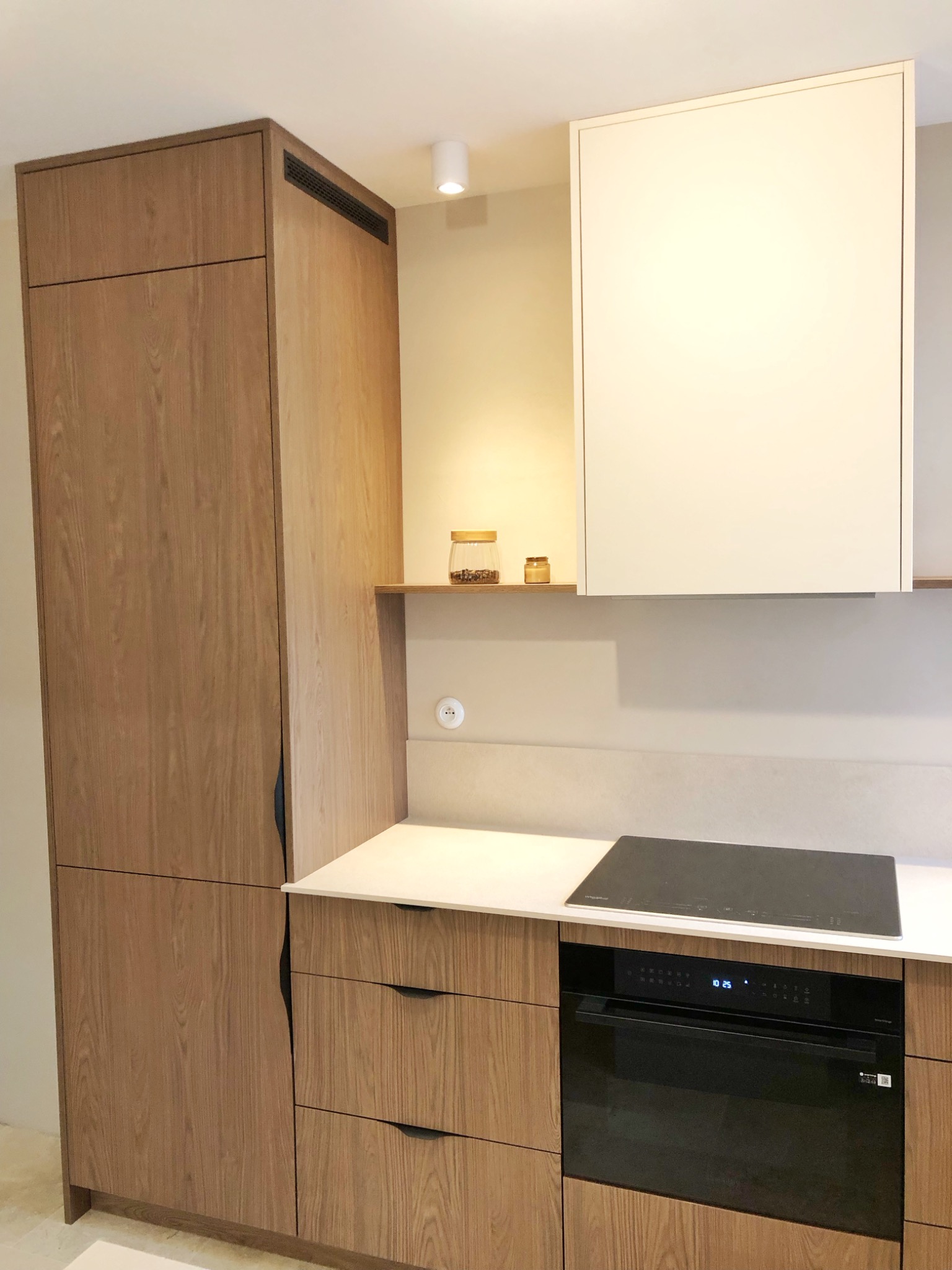 Nowoczesna zabudowa kuchenna z frontami z jasnego drewna, czarnym piekarnikiem i płytą indukcyjną, uzupełniona białymi szafkami wiszącymi i półką z dekoracyjnymi słoikami.