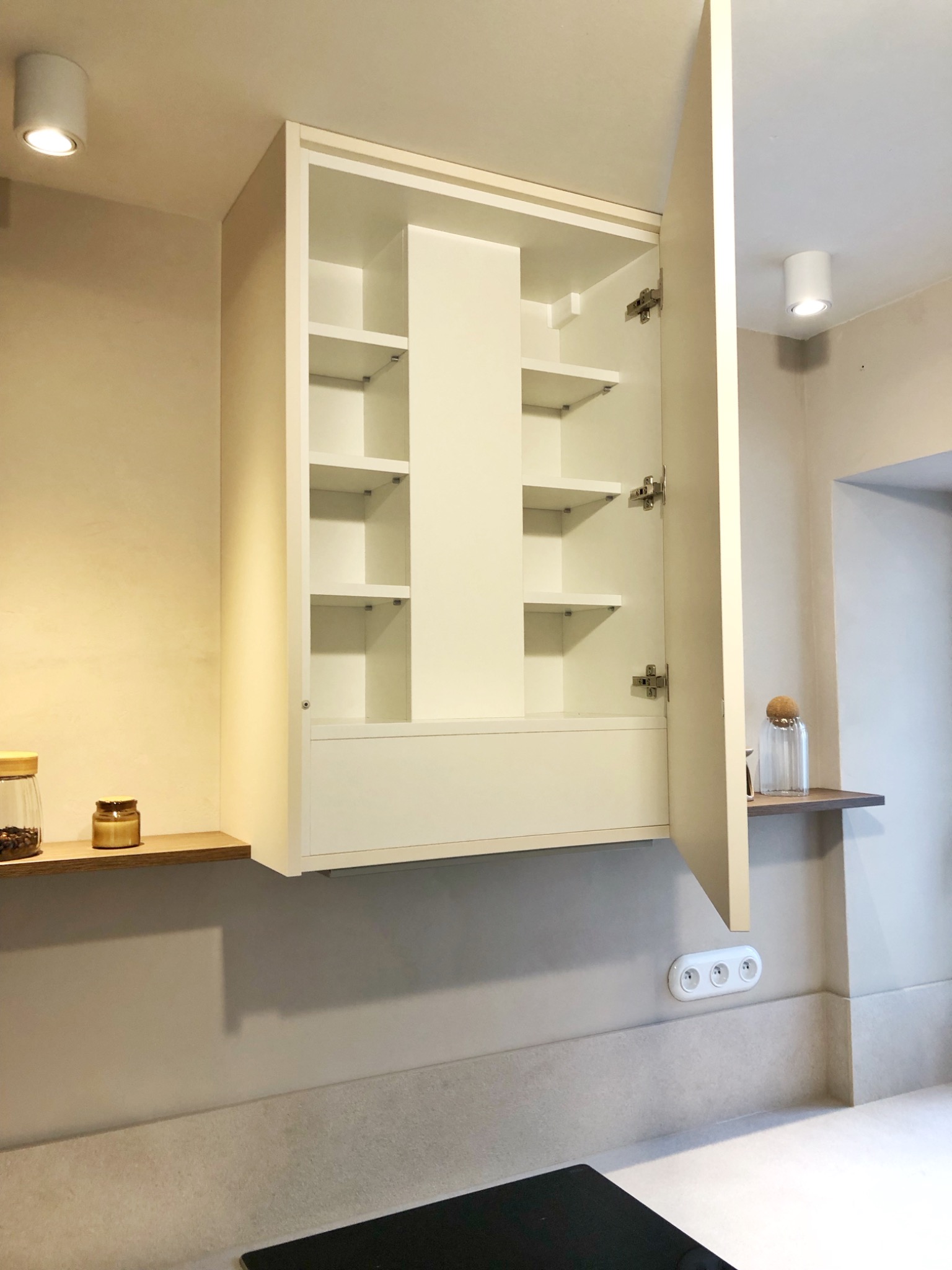 Biała szafka kuchenna wisząca z otwartymi drzwiami, ukazująca półki i środkowy pionowy panel, z drewnianą półką obok, na której stoją słoiki, oraz gniazdko elektryczne pod spodem.