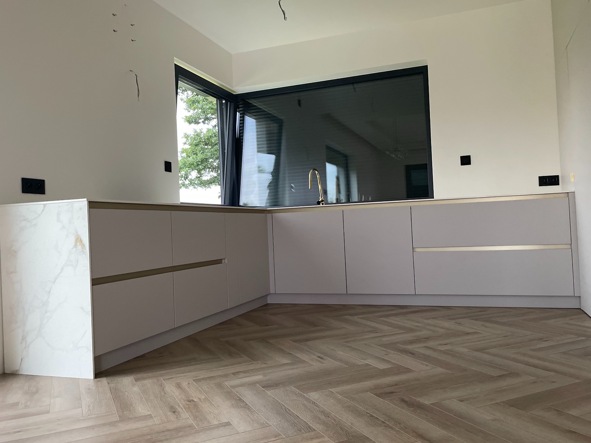 Kaszmirowa zabudowa kuchenna z poziomymi złotymi listwami, marmurowym panelem bocznym i złotą baterią przy oknie z widokiem na zieleń, ułożona na podłodze w jodełkę.