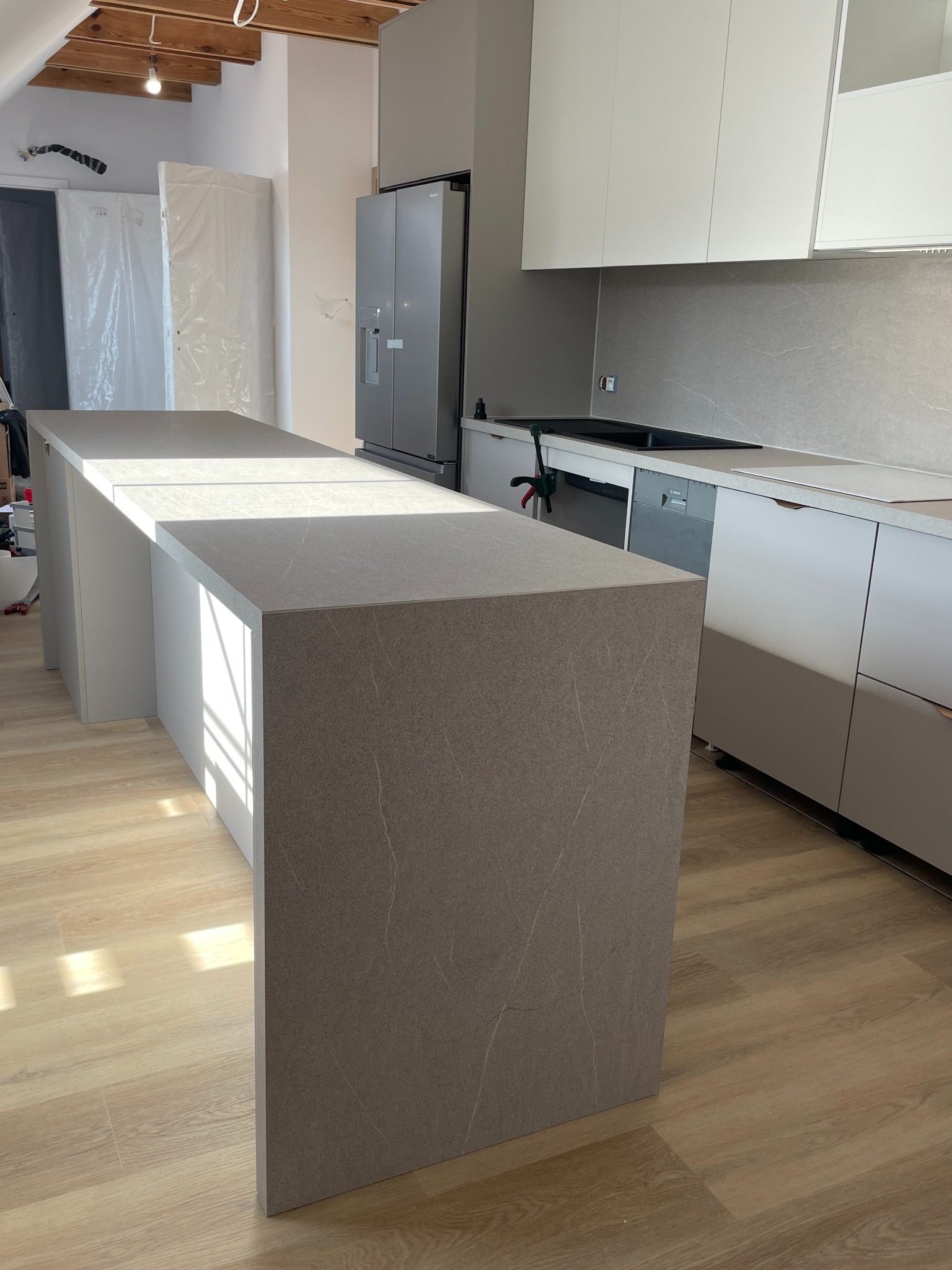 Nowoczesna kuchnia z szarym blatem wyspowym o marmurkowym wzorze, białymi szafkami i sprzętem AGD ze stali nierdzewnej; widoczne drewniane belki stropowe.