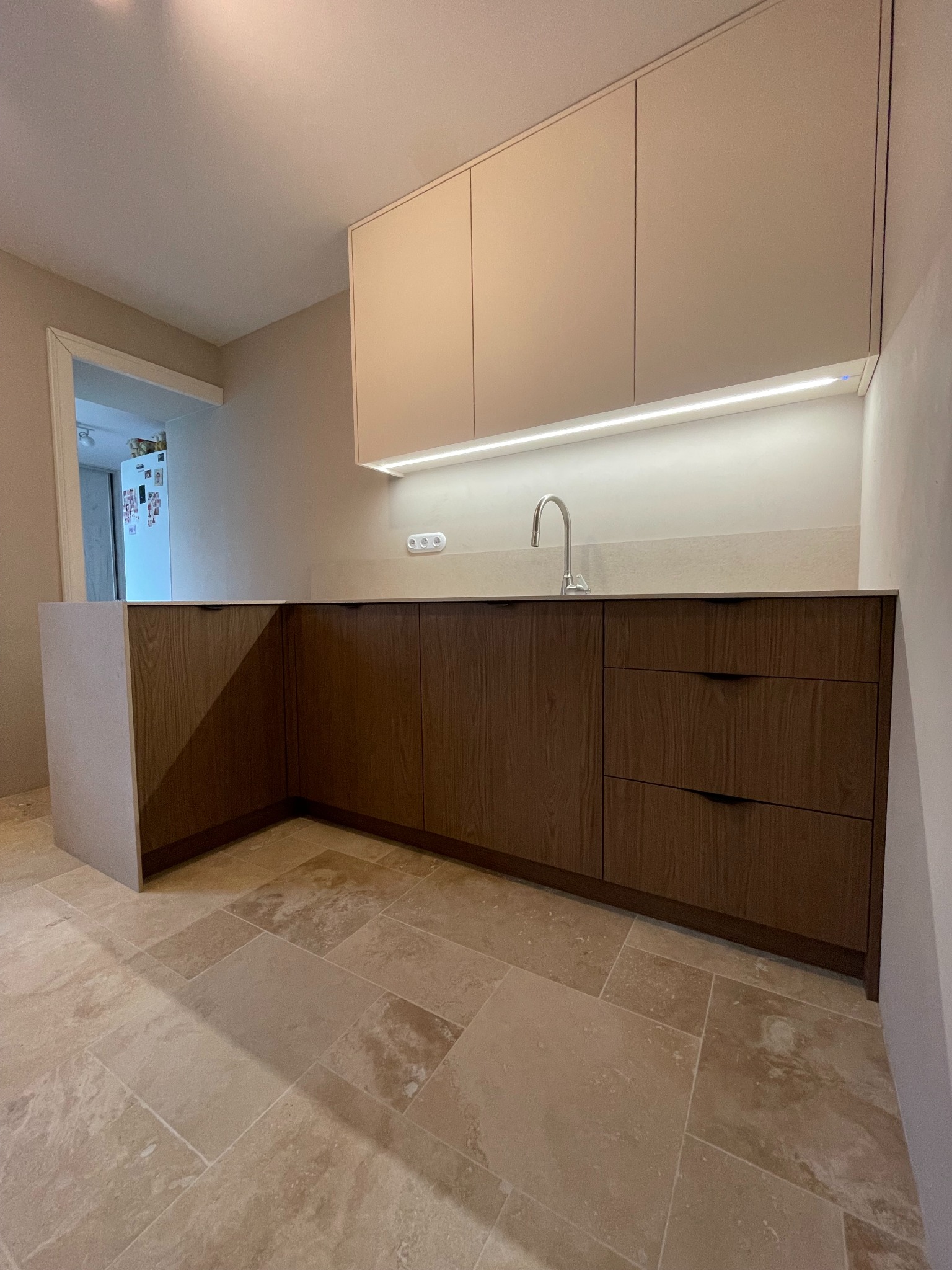 Nowoczesna zabudowa kuchenna z drewnianymi frontami i podświetleniem LED pod szafkami wiszącymi, widok na fragment blatu i baterię w minimalistycznym stylu, na podłodze płytki imitujące kamień.