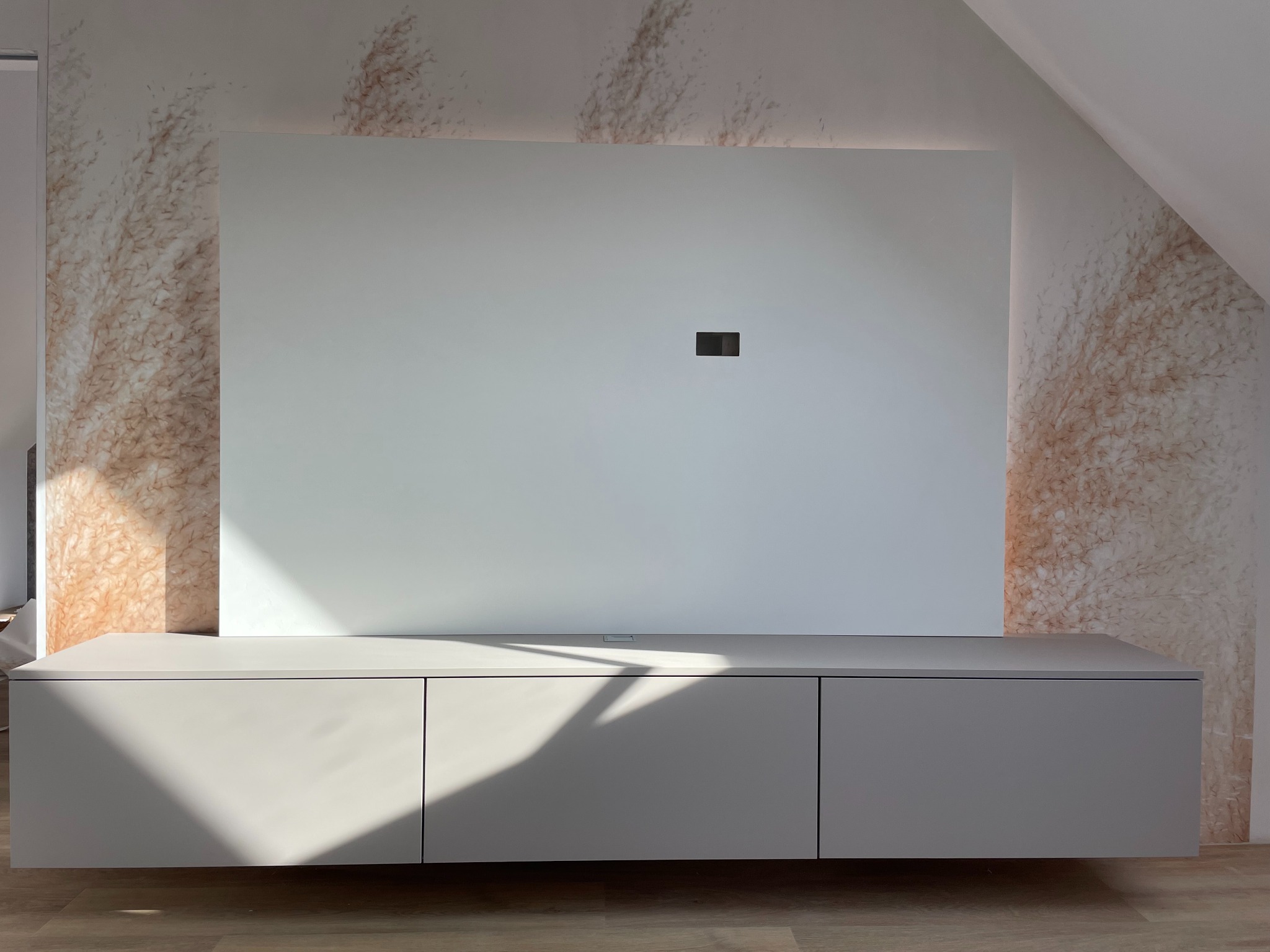 Szara szafka RTV podwieszana pod białą ścianką z ukrytym oświetleniem LED, na tle ściany z wzorem trawy pampasowej, w pokoju na poddaszu.