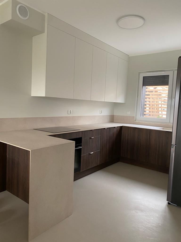 Nowoczesna kuchnia z białymi szafkami górnymi i ciemnobrązowymi szafkami dolnymi, blatem w kolorze beżowym, widok na okno z żaluzjami.