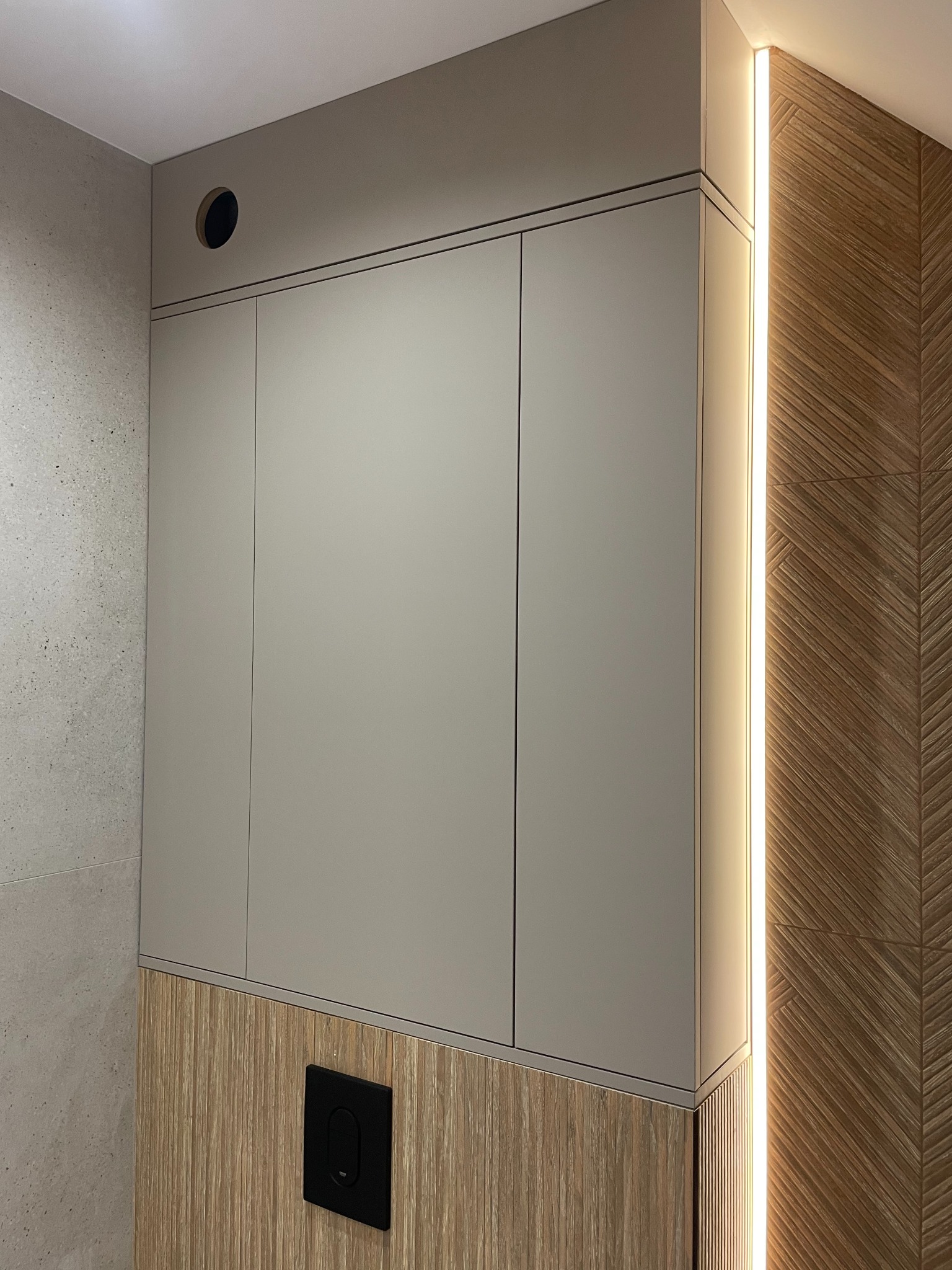 Beżowa szafka wisząca z trzema drzwiczkami, zamontowana w łazience z drewnianą boazerią i oświetleniem LED, widoczny przycisk spłuczki.