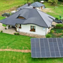 Dom jednorodzinny z instalacją fotowoltaiczną na gruncie, widok z góry, zielony trawnik, szara dachówka, okno dachowe w kształcie półksiężyca.