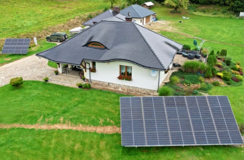 Dom jednorodzinny z instalacją fotowoltaiczną na gruncie, widok z góry, zielony trawnik, szara dachówka, okno dachowe w kształcie półksiężyca.