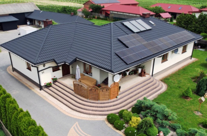 Dom jednorodzinny z ciemną dachówką i panelami fotowoltaicznymi na dachu, widok z góry, zadbany ogród z tujami i kostką brukową.