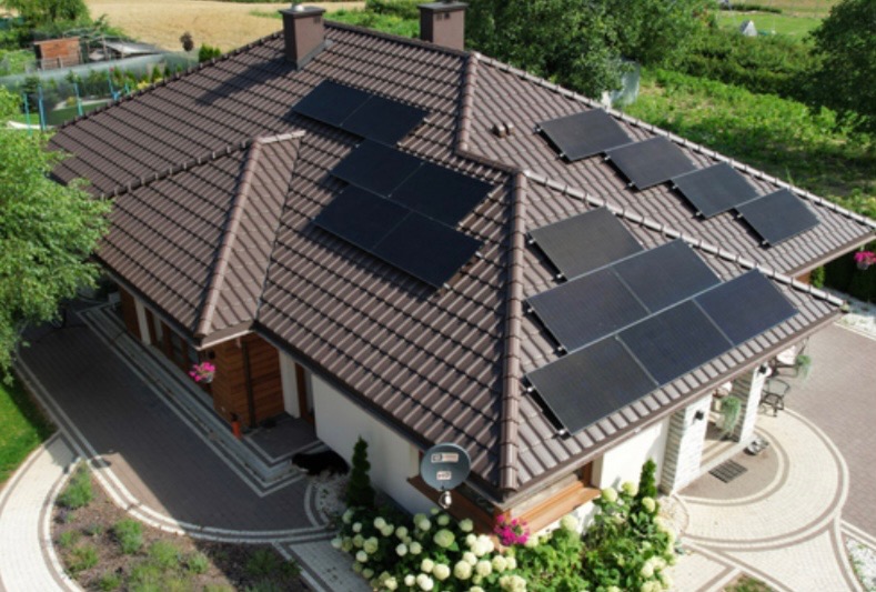 Dom jednorodzinny z brązowym dachem pokrytym panelami słonecznymi, widok z góry, starannie zagospodarowany ogród z kostką brukową i roślinnością.