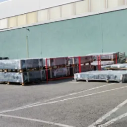 Gotowe elementy konstrukcji stalowej, zabezpieczone folią, ułożone na paletach na placu budowy, w tle ściana hali w kolorze seledynowym.