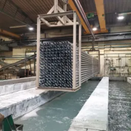 Metalowe rury zanurzane w zbiorniku z płynem podczas procesu obróbki powierzchniowej w hali przemysłowej.