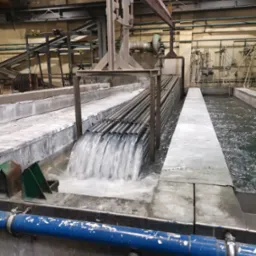 Rury metalowe zawieszone na suwnicy zanurzane w zbiorniku z wodą podczas procesu płukania w hali przemysłowej.