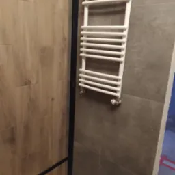 Fragment łazienki z grzejnikiem drabinkowym, płytkami imitującymi drewno i szarą ścianą z widocznym fragmentem kabiny prysznicowej.