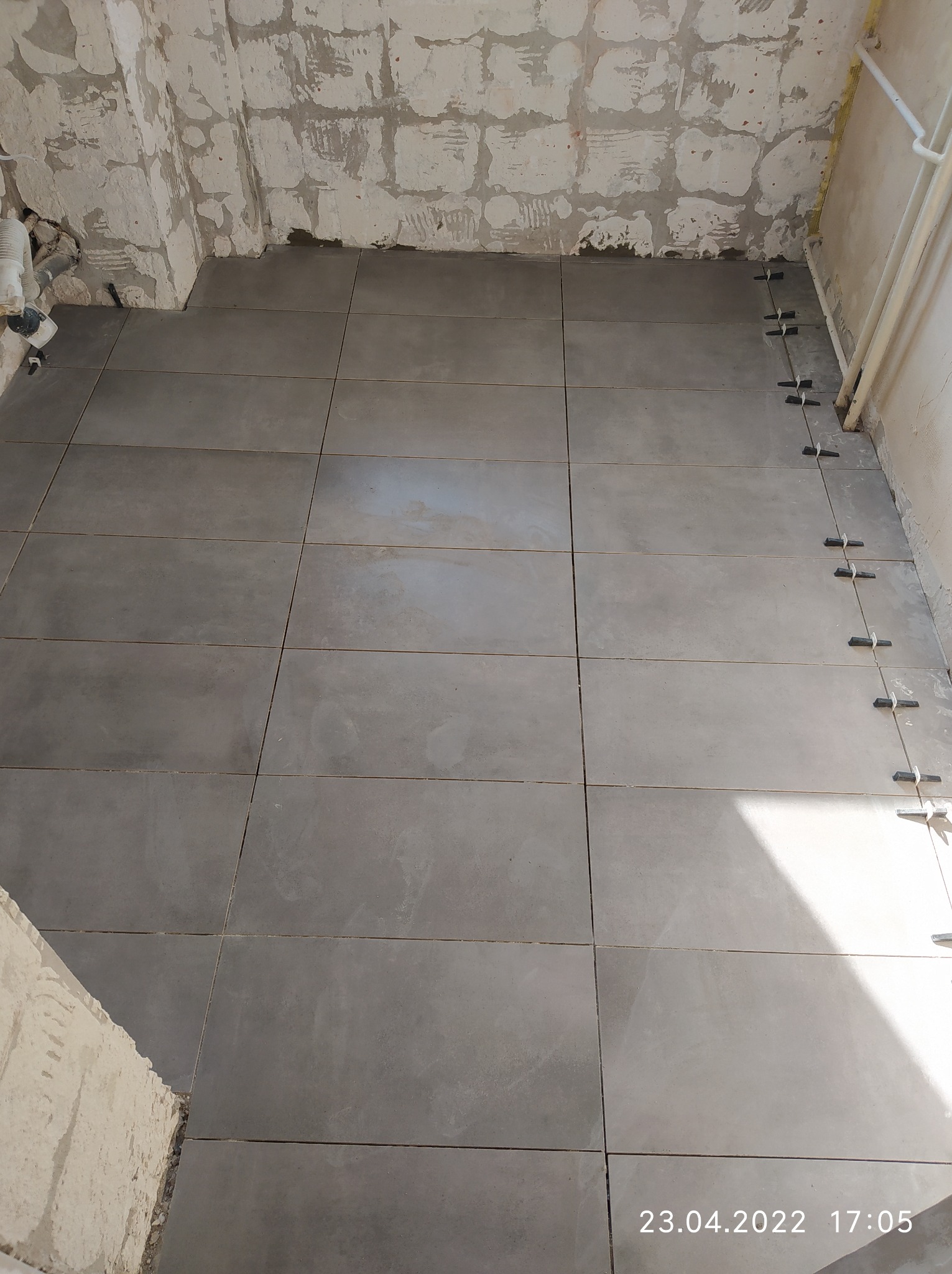 Szare płytki podłogowe ułożone w łazience w trakcie remontu, widoczne krzyżyki dystansowe, ściany surowe, przygotowane do dalszych prac wykończeniowych.