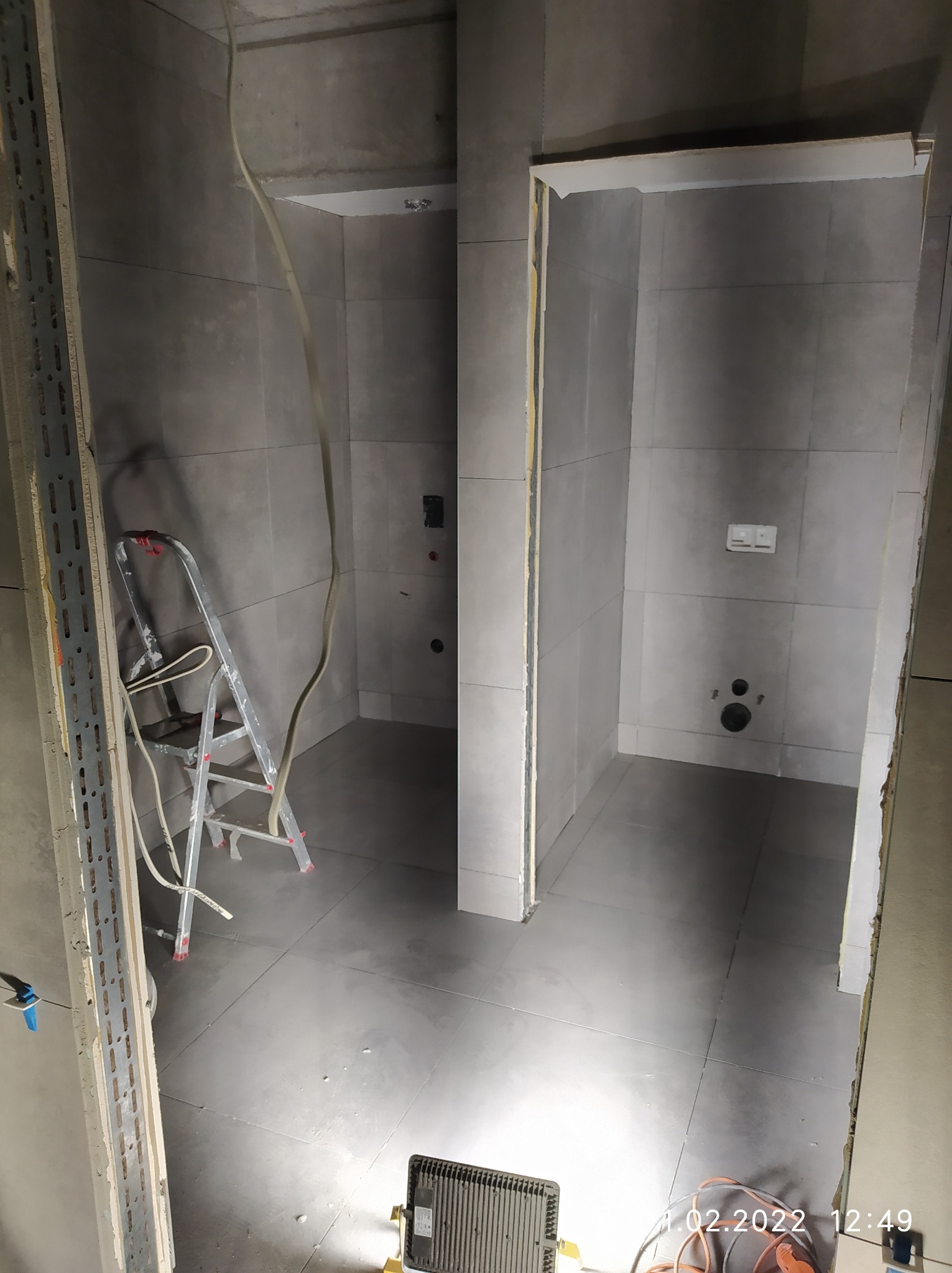 Wnętrze łazienki w trakcie prac wykończeniowych: szare płytki na ścianach i podłodze, widoczna drabina, przewody elektryczne, metalowy profil, oświetlenie robocze.