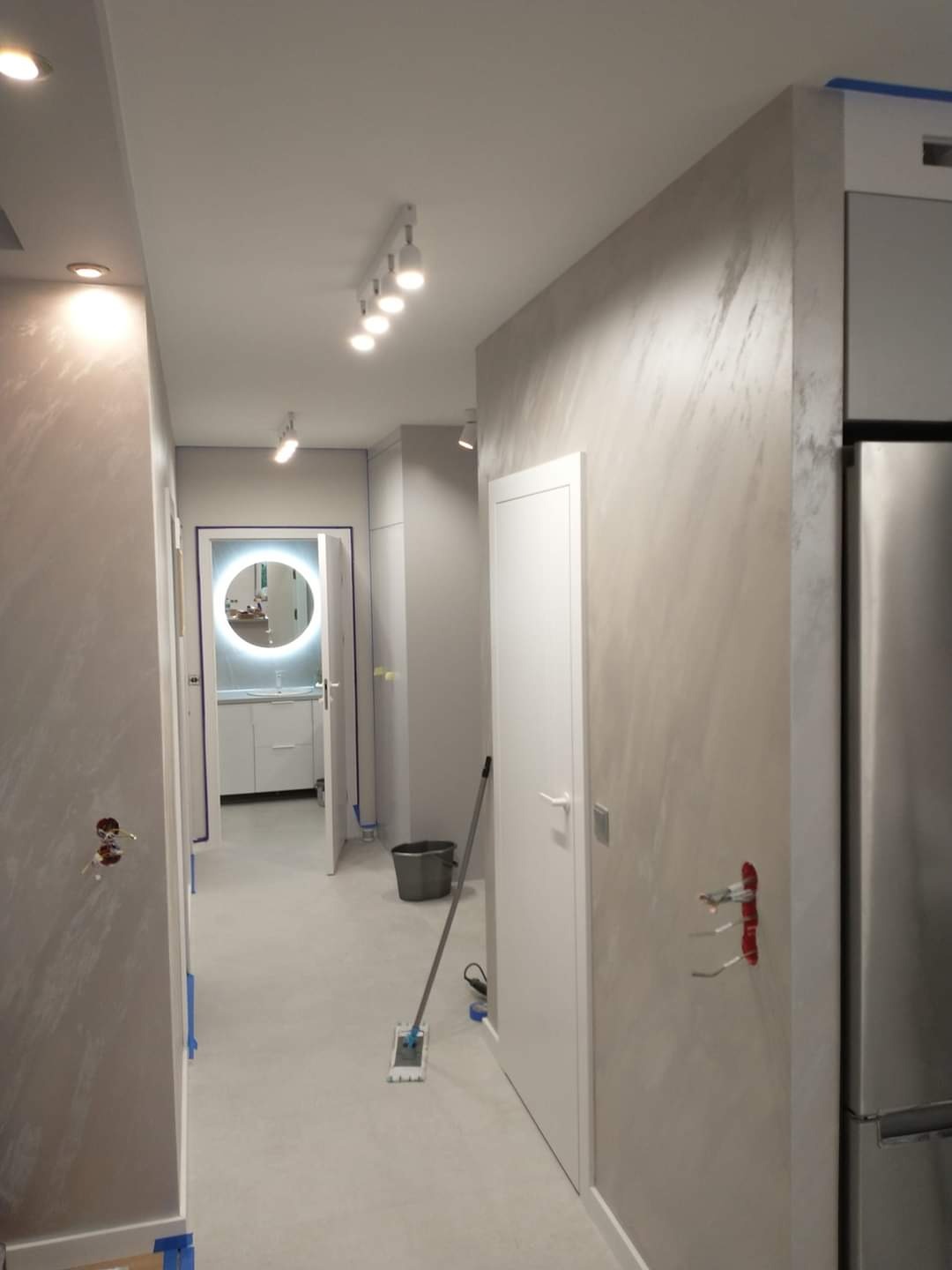 Korytarz po świeżym malowaniu z efektem dekoracyjnym na ścianach, widoczna łazienka z okrągłym lustrem z podświetleniem, mop na podłodze i otwarte puszki elektryczne.