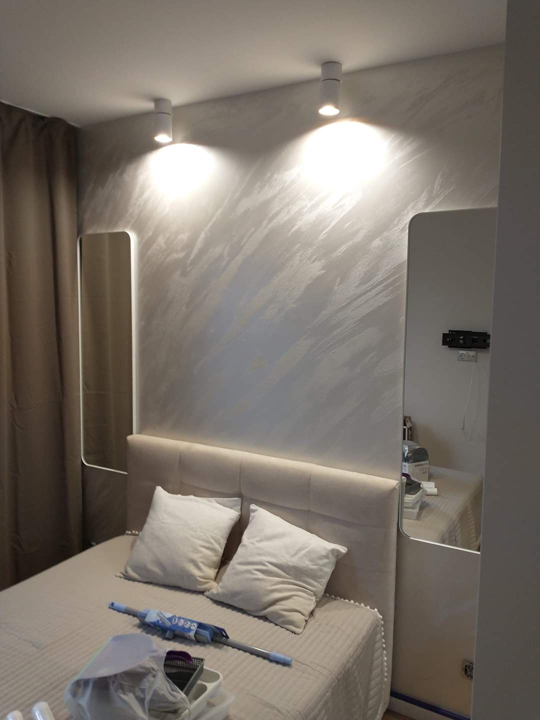 Ściana w sypialni z dekoracyjną, srebrzystą fakturą, oświetlona dwoma punktowymi lampami sufitowymi, z lustrami po bokach łóżka i widocznymi narzędziami malarskimi na łóżku.