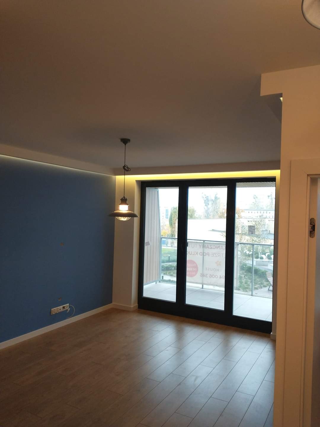 Pomalowane na niebiesko wnętrze pokoju z ułożonymi panelami podłogowymi i dużym oknem balkonowym z czarną ramą, oświetlone wiszącą lampą i listwą LED pod sufitem.