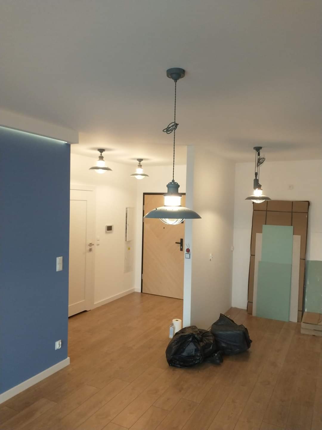 Wnętrze mieszkania po odświeżeniu, z nową, błękitną ścianą, drewnianą podłogą i trzema lampami w stylu industrialnym. Na podłodze leżą worki na śmieci, a w tle widoczne są płyty gipsowo-kartonowe.