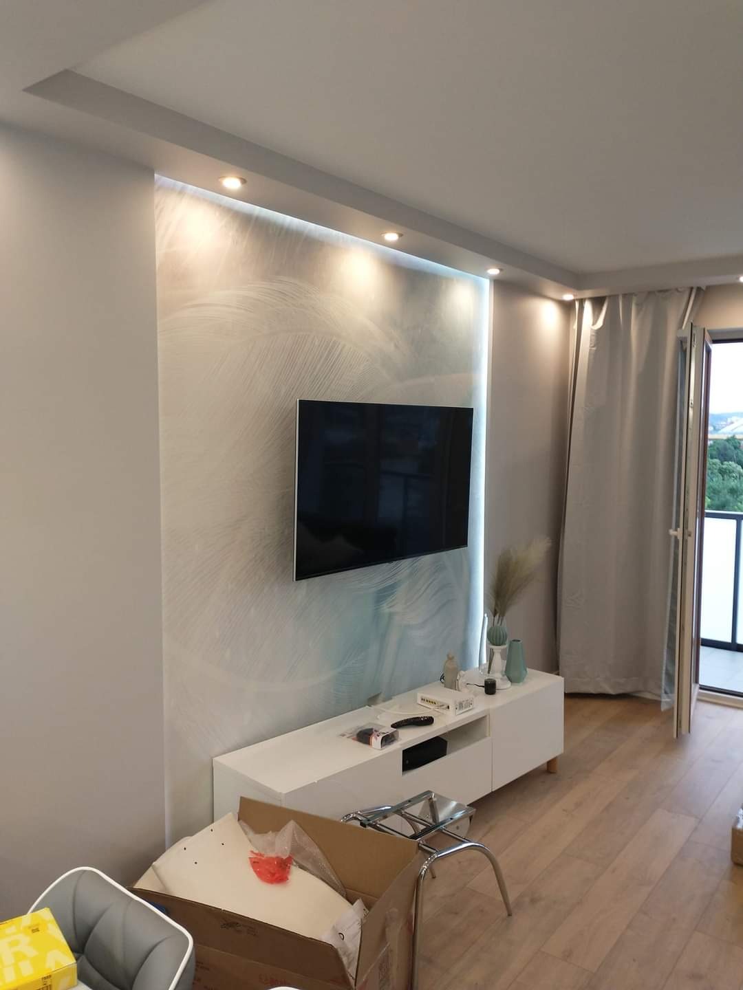 Telewizor zawieszony na ścianie z dekoracyjną tapetą w odcieniach szarości i błękitu, podświetloną punktowymi lampami LED, biała szafka RTV z dekoracjami, drewniana podłoga, częściowo widoczne...