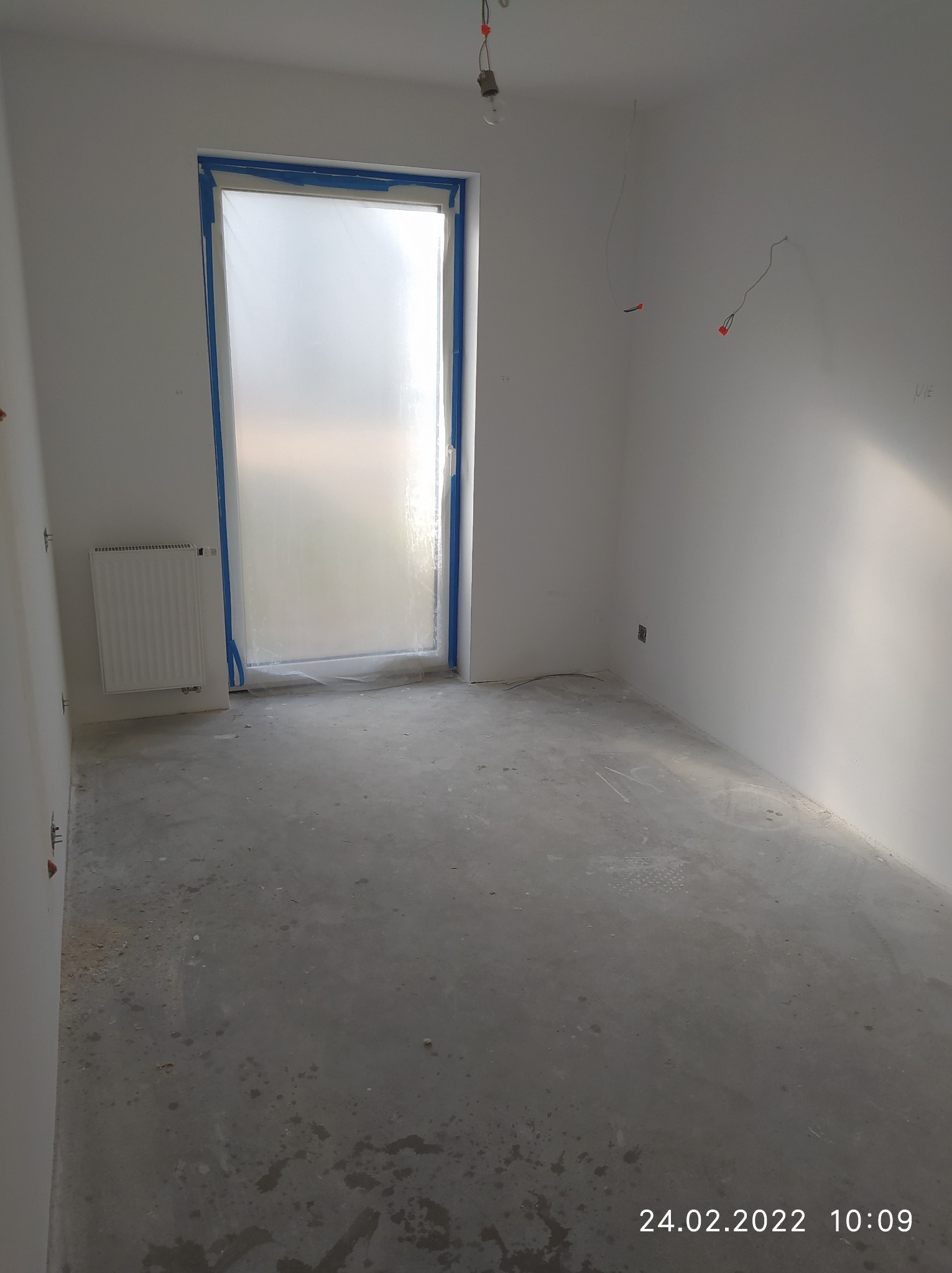 Pokój w trakcie przygotowań do prac wykończeniowych: zabezpieczone okno folią i taśmą malarską, surowy beton na podłodze, ściany przygotowane do malowania, widoczne wystające kable elektryczne.