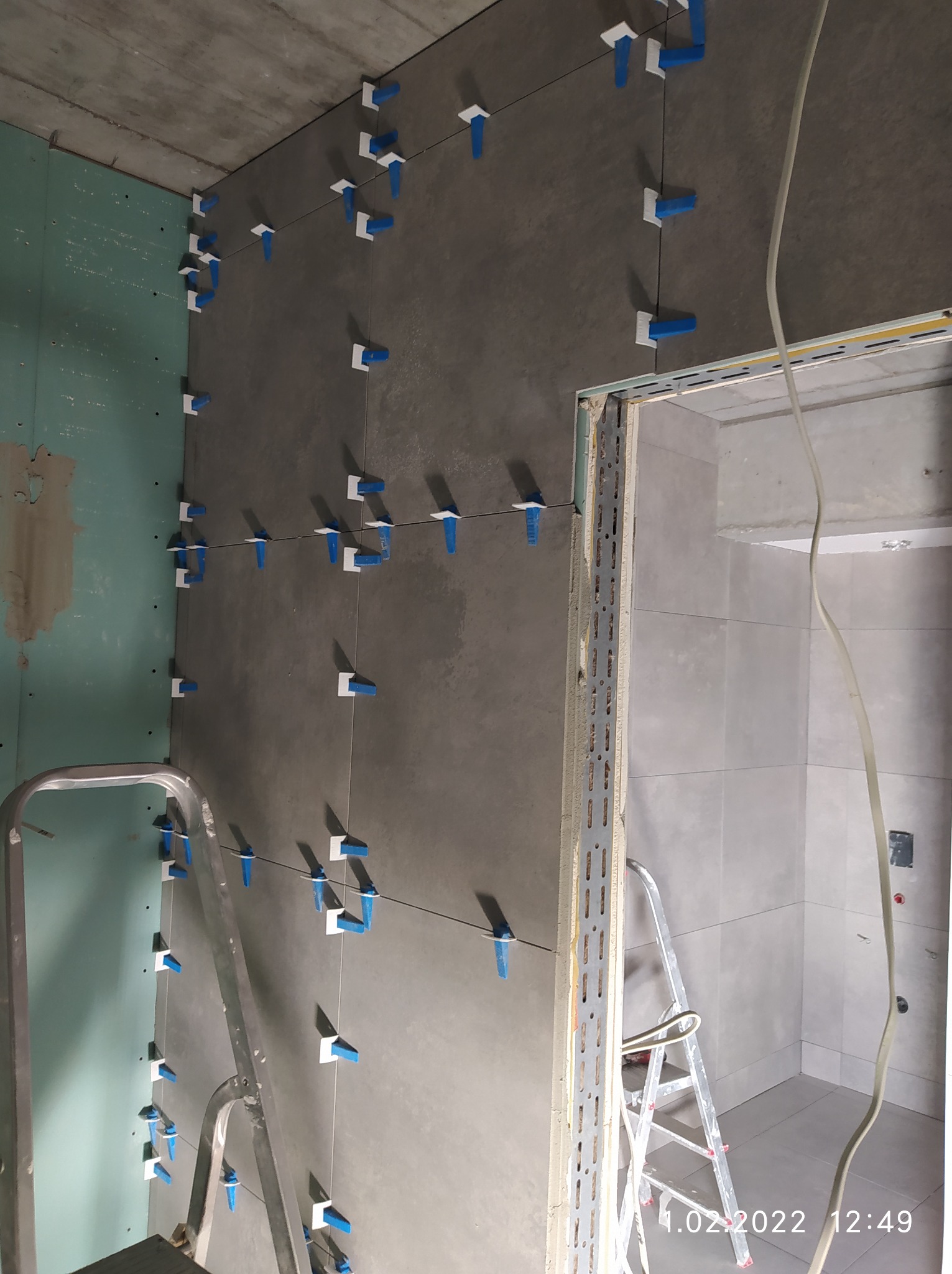 Szare płytki ścienne z systemem poziomowania płytek (białe podstawki i niebieskie kliny) w trakcie montażu, widoczne drabiny i surowy stan wykończenia wnętrza.