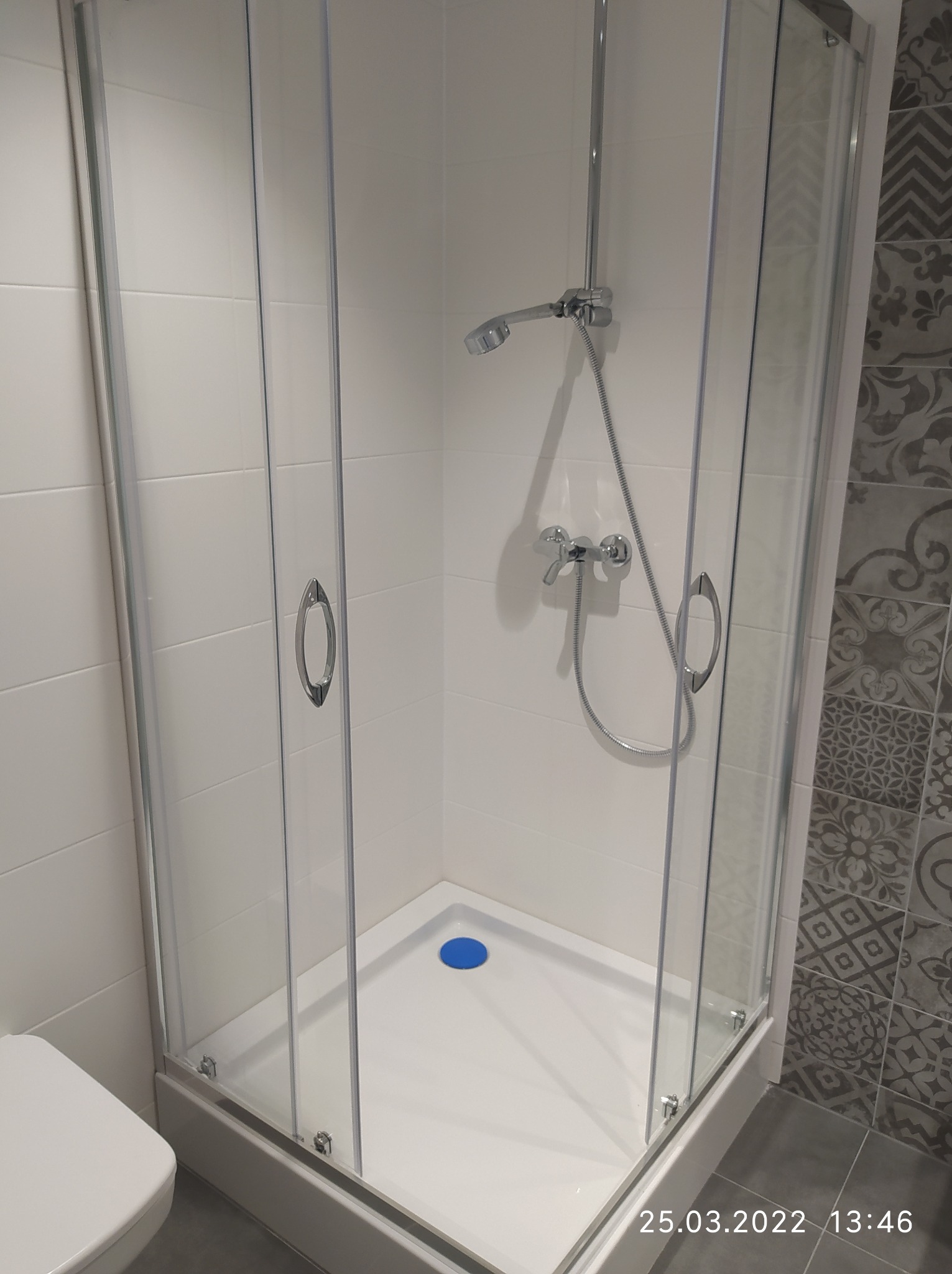 Kabina prysznicowa z chromowanymi elementami i deszczownicą, białe płytki ceramiczne na ścianach, dekoracyjna płytka wzorzysta w odcieniach szarości po prawej stronie, niebieski odpływ liniowy...