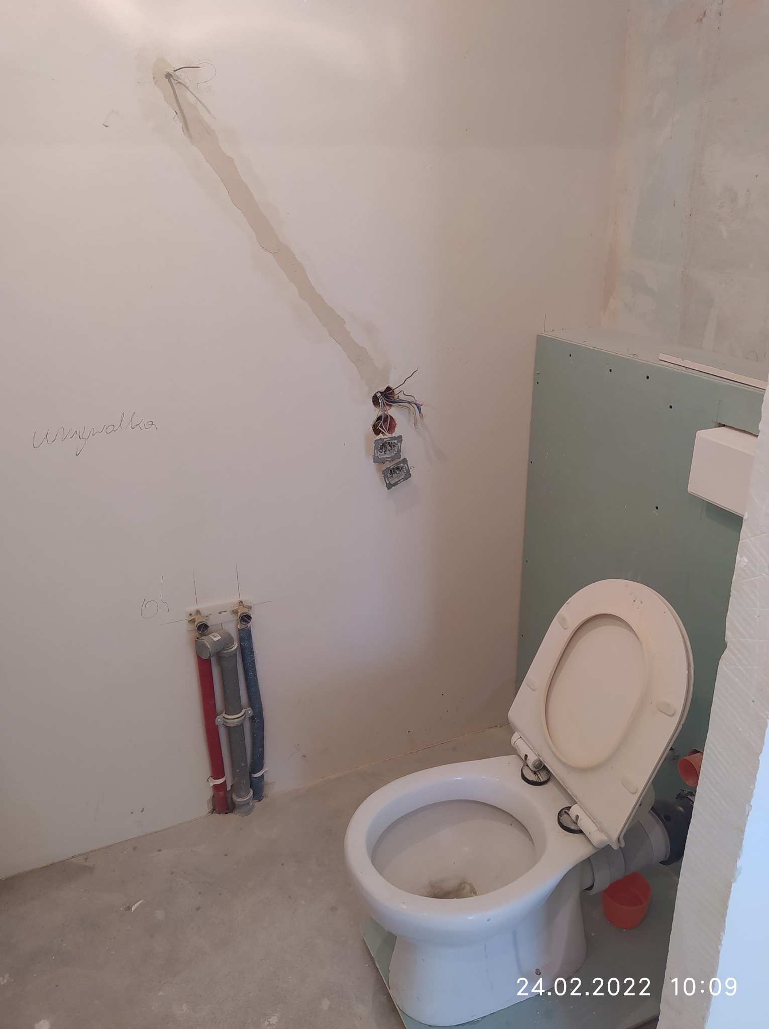 Niedokończona toaleta w trakcie instalacji: widoczne rury, przewody elektryczne, płyta kartonowo-gipsowa i napis 'umywalka' na ścianie.