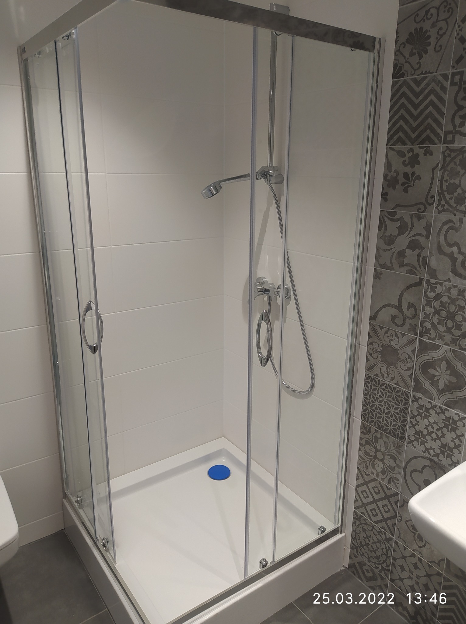 Nowoczesna kabina prysznicowa z chromowanymi elementami, biały brodzik z niebieskim odpływem, ściana wyłożona białymi płytkami oraz dekoracyjnymi płytkami w odcieniach szarości.