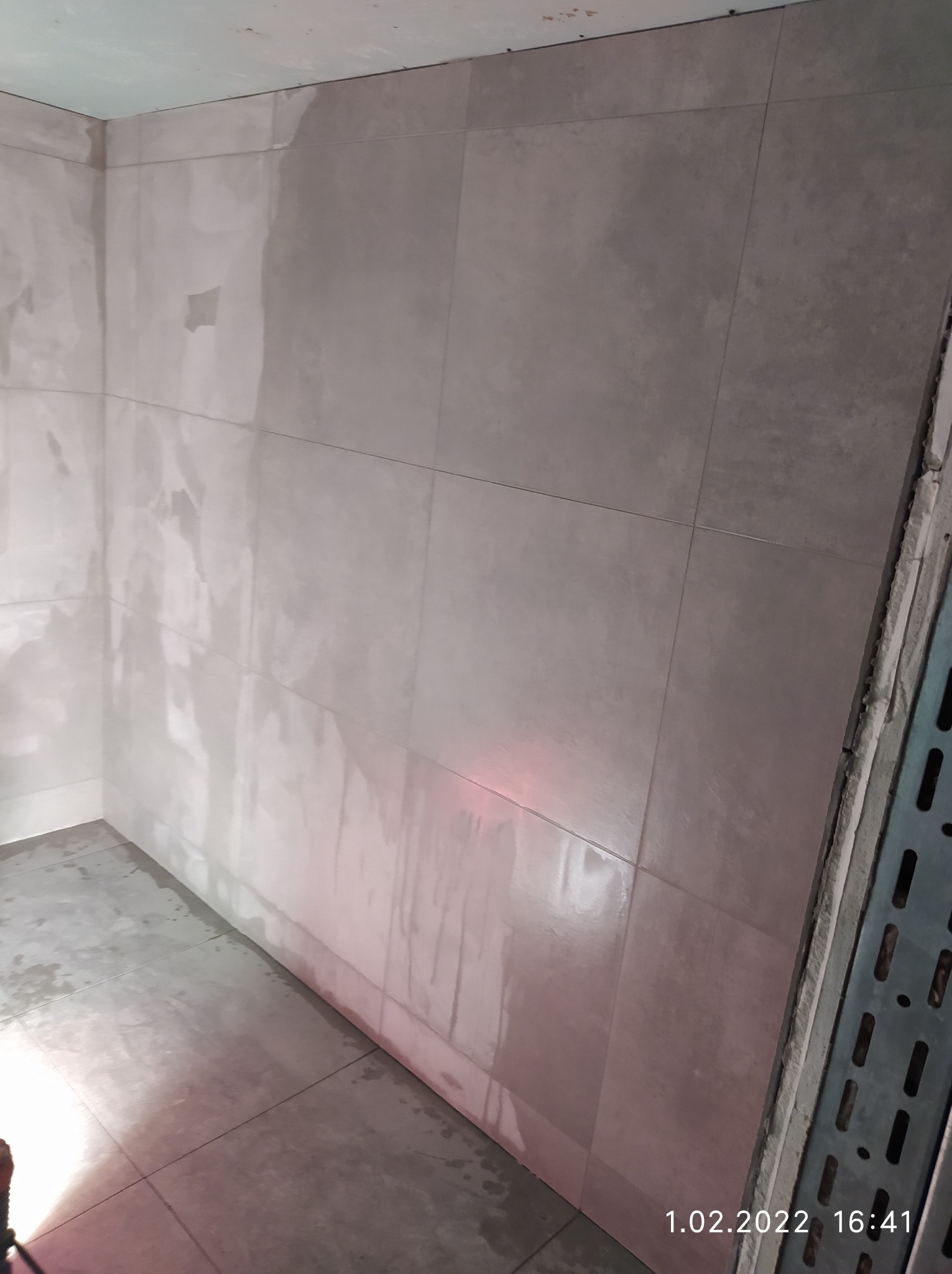 Świeżo ułożone, duże, szare płytki podłogowe i ścienne w łazience, jeszcze mokre od kleju, z widocznymi śladami po czyszczeniu i metalową listwą przypodłogową w prawym rogu.