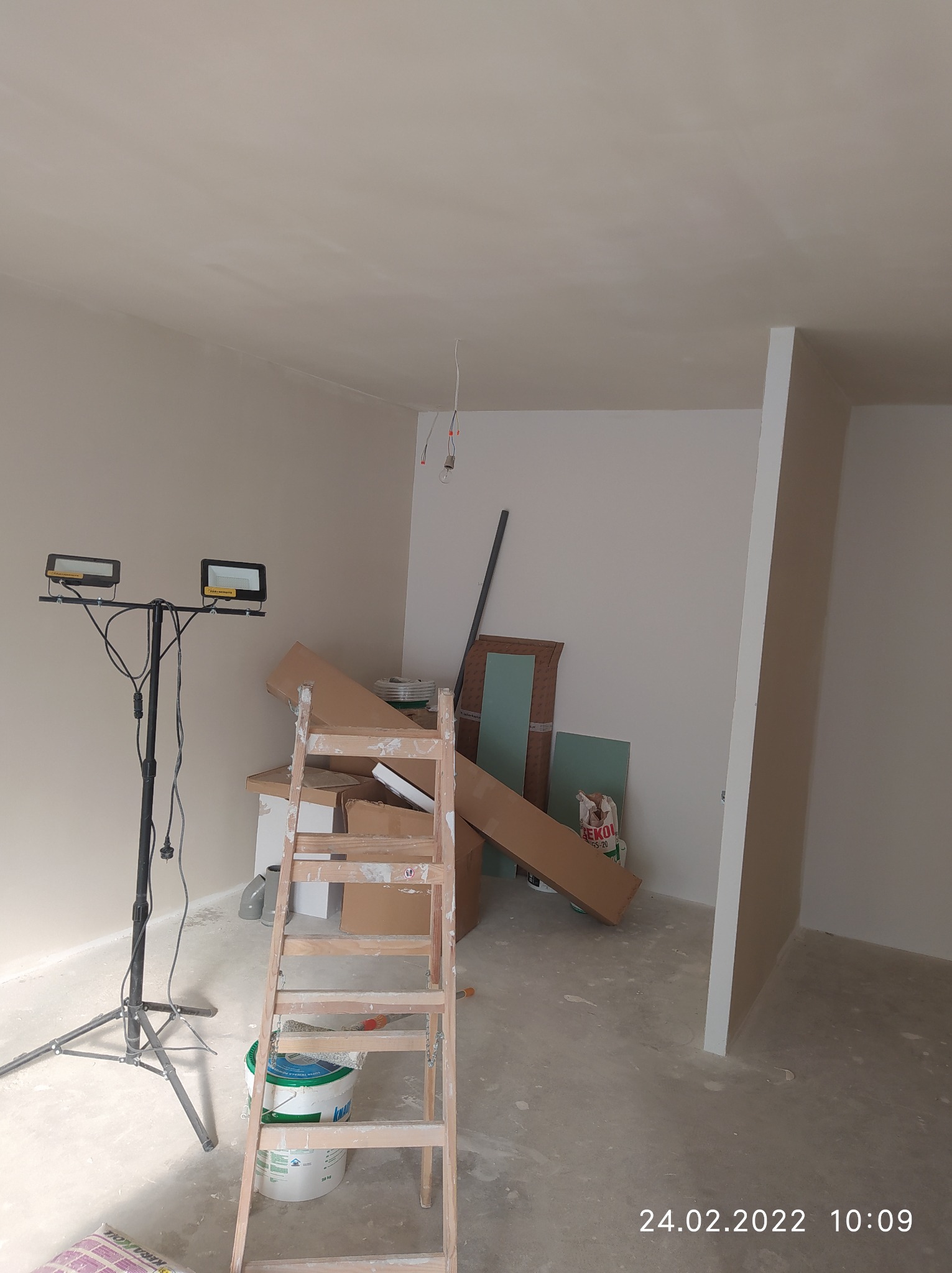 Wnętrze w trakcie prac wykończeniowych, widoczna drabina oparta o ścianę, puszka z farbą, kartony, lampa budowlana na statywie i częściowo wygładzona ściana oraz sufit.