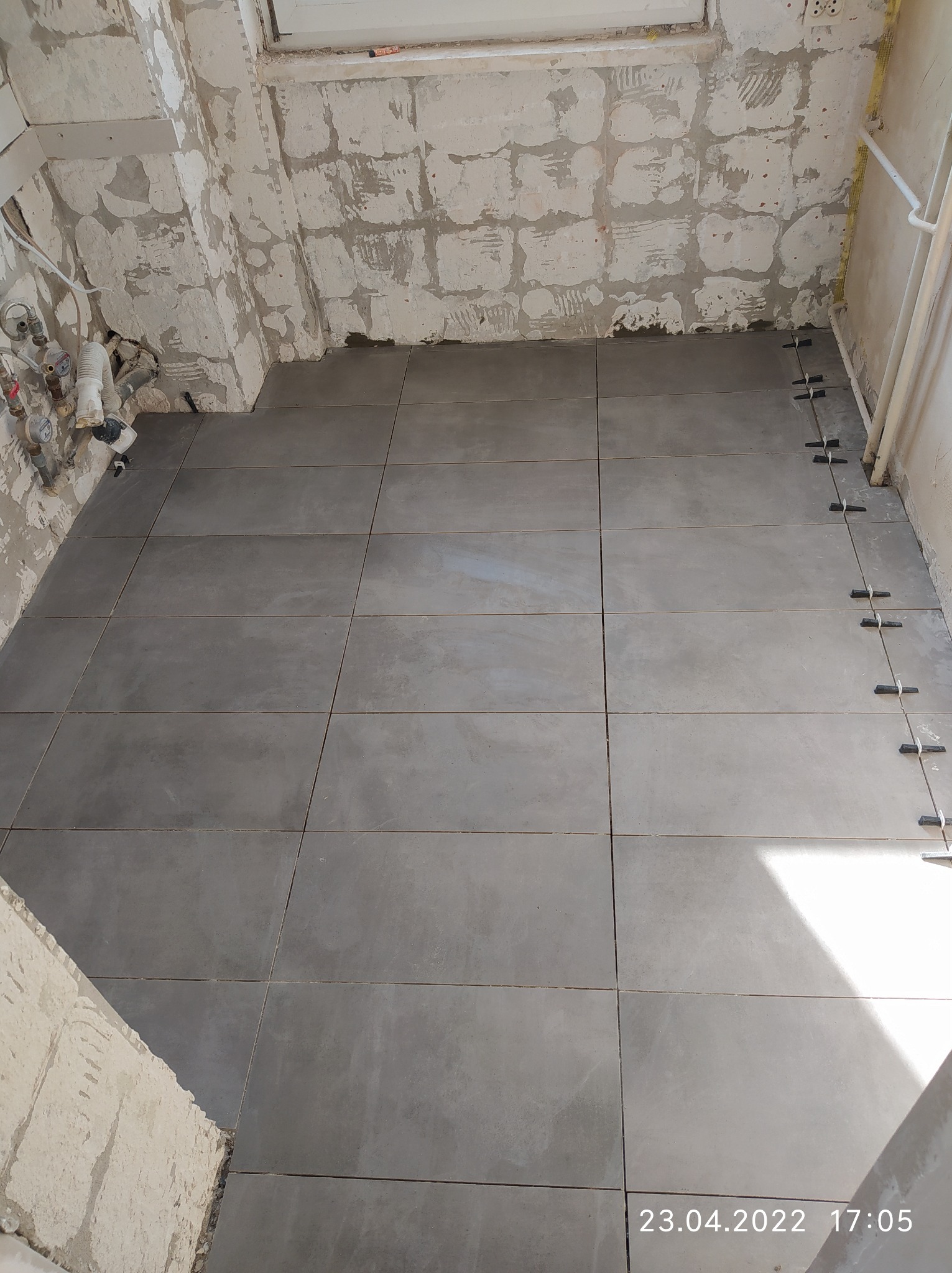 Szare płytki podłogowe w trakcie układania w łazience, widoczne krzyżyki dystansowe, surowe ściany przygotowane do dalszych prac wykończeniowych, instalacja wodna i okno w tle.