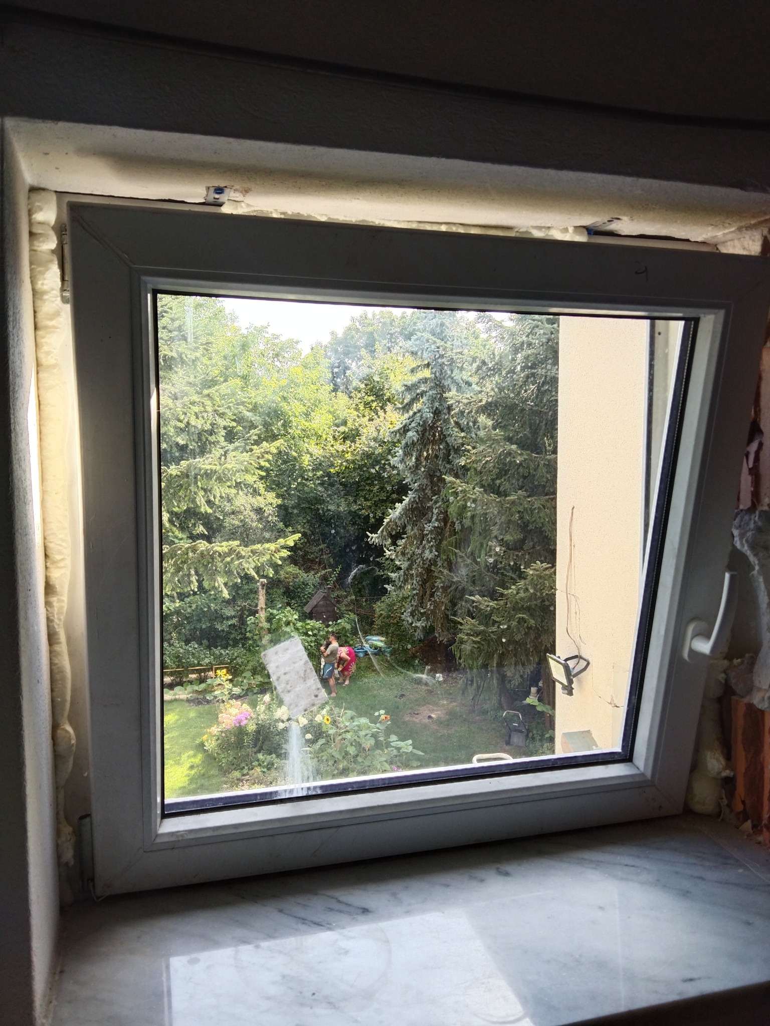 Nowe okno PCV z widoczną pianką montażową uszczelniającą ramę, widok na ogród z drzewami i kwiatami.